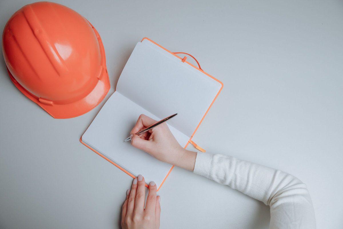 Hand writing in a notebook beside an orange hard hat